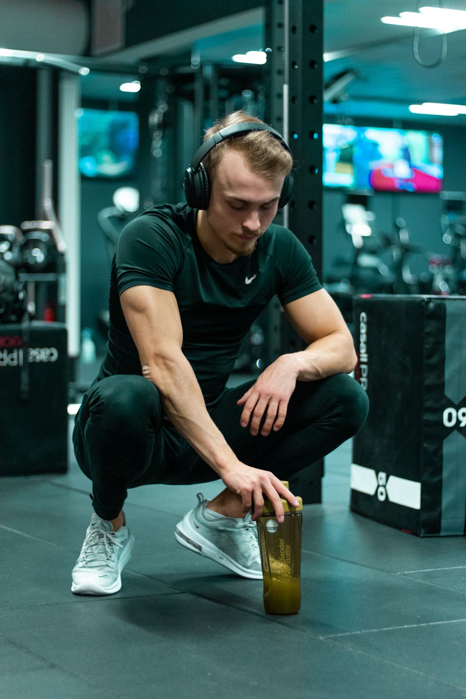 Ein Mann in Sportkleidung hockt im Fitnessstudio und hält ein grünes Getränk.
