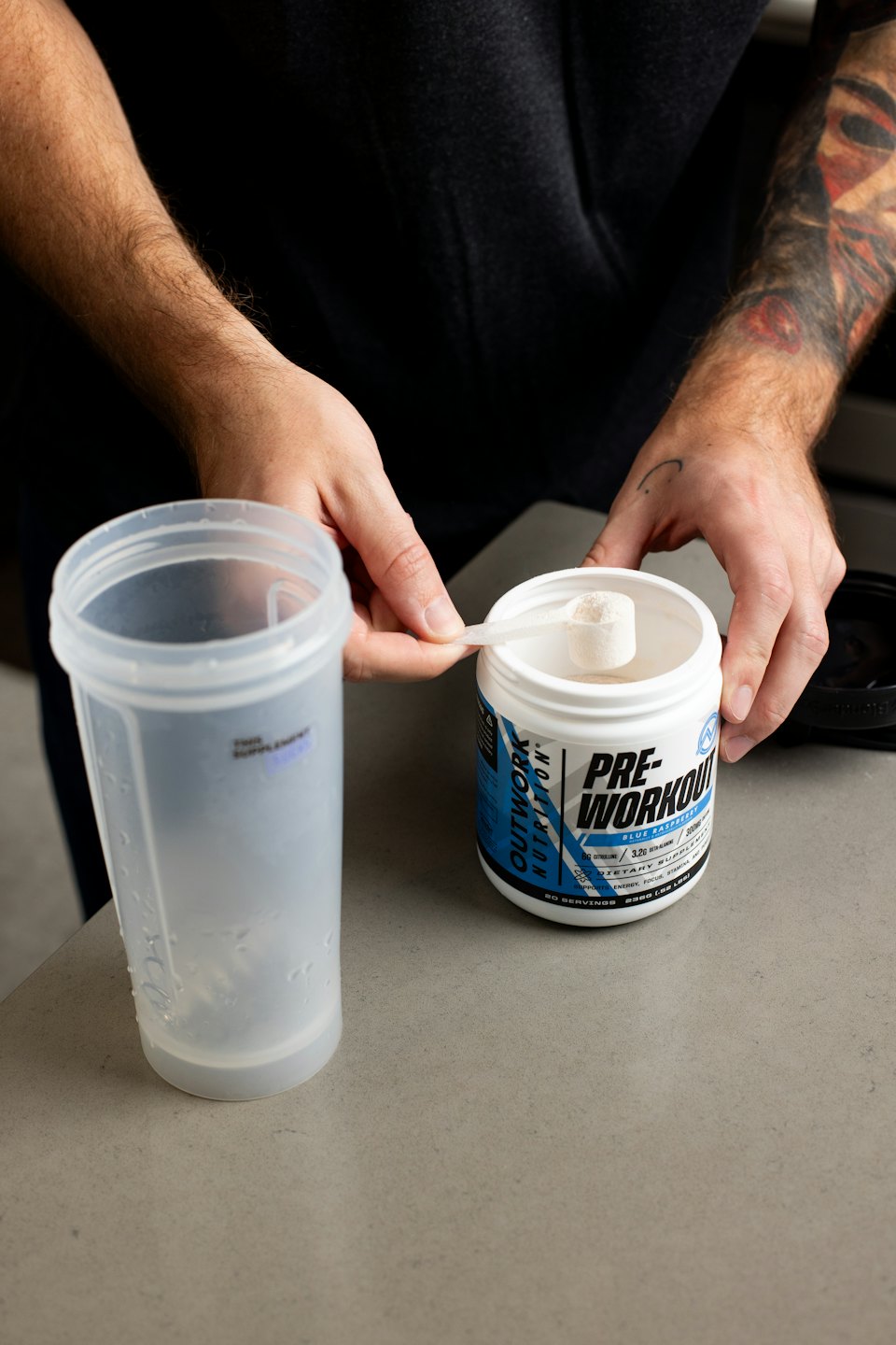 Person bereitet Pre-Workout-Supplement mit Schaufel und Shaker-Flasche zu.